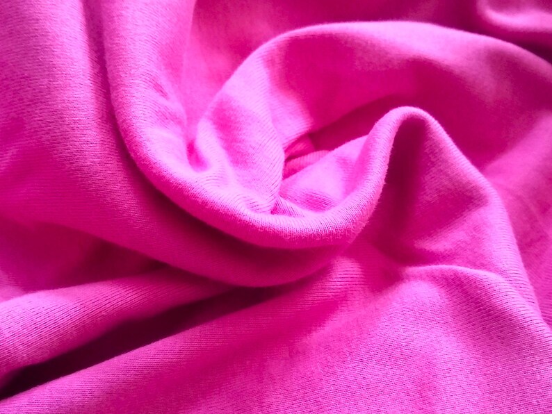 May include: Close-up of a vibrant fuchsia fabric with soft folds and subtle textures. The material appears to be a knit or jersey, with a smooth, slightly shiny surface. The image highlights the fabric's color and drape.