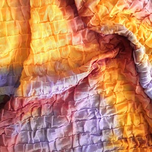 Może przedstawiać: Zbliżenie na tkaninę tie-dye z fakturowaną, pomarszczoną powierzchnią. Tkanina jest połączeniem kolor&oacute;w pomarańczowego, ż&oacute;łtego, fioletowego i r&oacute;żowego.