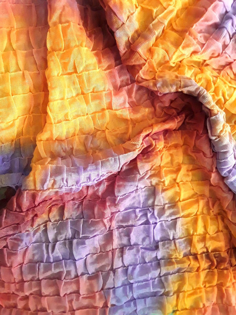Może przedstawiać: Zbliżenie na tkaninę tie-dye z fakturowaną, pomarszczoną powierzchnią. Tkanina jest połączeniem kolor&oacute;w pomarańczowego, ż&oacute;łtego, fioletowego i r&oacute;żowego.