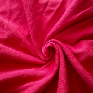 May include: Close-up of a vibrant fuchsia fabric, tightly swirled to create a spiral effect. The soft material has a textured surface, with visible folds and shadows that enhance the depth and dimension of the textile. The bright color dominates the frame.
