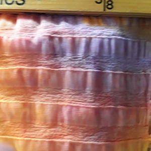 Może przedstawiać: Zbliżenie na tkaninę z wzorem tie-dye w odcieniach pomarańczowego, r&oacute;żowego i fioletowego. Tkanina ma fakturowaną, pomarszczoną strukturę. Na g&oacute;rze obrazu widoczna jest linijka z zaznaczonym pomiarem "5/8".