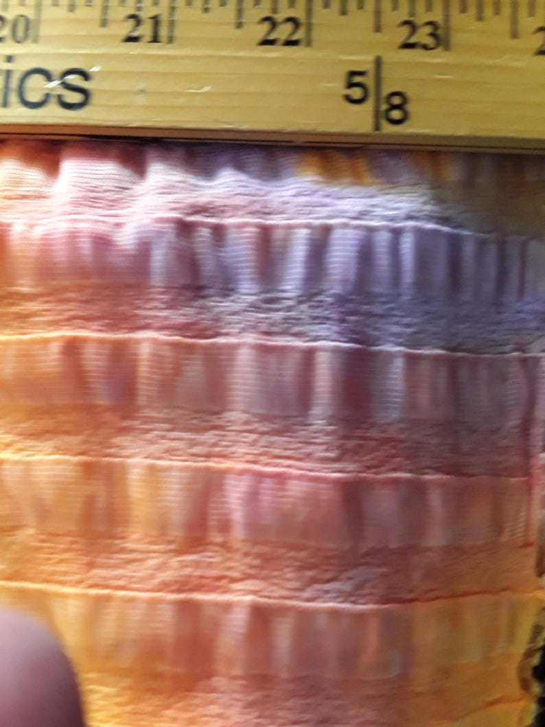 Może przedstawiać: Zbliżenie na tkaninę z wzorem tie-dye w odcieniach pomarańczowego, r&oacute;żowego i fioletowego. Tkanina ma fakturowaną, pomarszczoną strukturę. Na g&oacute;rze obrazu widoczna jest linijka z zaznaczonym pomiarem "5/8".