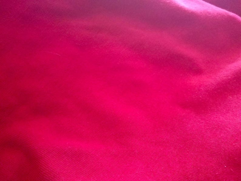 May include: A close-up shot of a vibrant, fuchsia-colored fabric. The material has a soft, slightly textured appearance, with subtle variations in tone creating depth. The fabric is smooth and appears to be draped, with gentle folds and shadows.