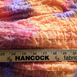 Może przedstawiać: Zbliżenie na tkaninę tie-dye o pomarszczonej fakturze. Tkanina jest w kolorze pomarańczowym, r&oacute;żowym i fioletowym. Na dole obrazu widoczny jest drewniany linijka z napisem "Hancock Fabrics".