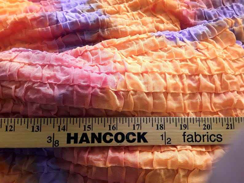 Może przedstawiać: Zbliżenie na tkaninę tie-dye o pomarszczonej fakturze. Tkanina jest w kolorze pomarańczowym, r&oacute;żowym i fioletowym. Na dole obrazu widoczny jest drewniany linijka z napisem "Hancock Fabrics".