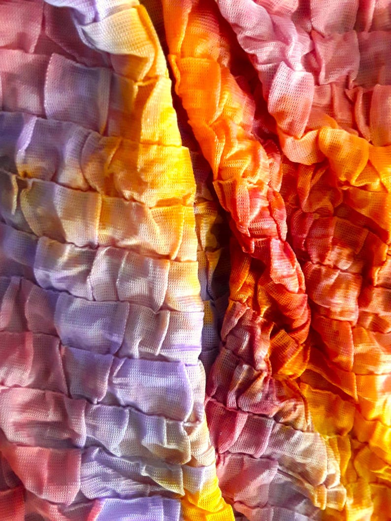 Może przedstawiać: Zbliżenie na tkaninę z wzorem tie-dye w odcieniach fioletu, pomarańczy i ż&oacute;łci. Tkanina jest fakturowana i ma pomarszczony wygląd.