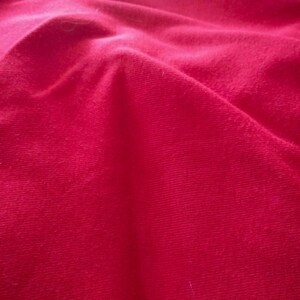 May include: Close-up of a vibrant red fabric with soft folds and a textured surface. The material appears to be a plush textile, possibly velvet or a similar material, with subtle variations in color.
