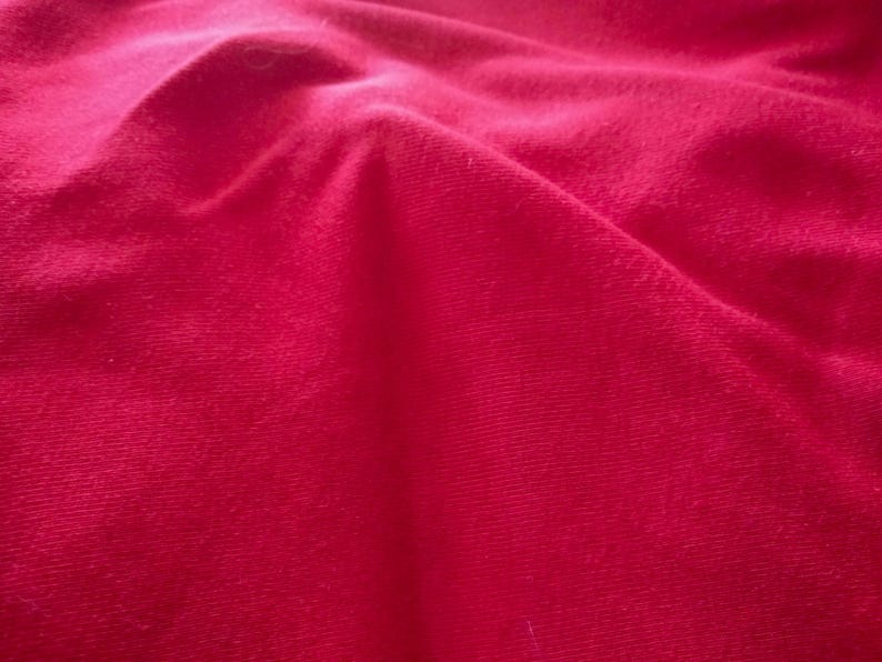 May include: Close-up of a vibrant red fabric with soft folds and a textured surface. The material appears to be a plush textile, possibly velvet or a similar material, with subtle variations in color.