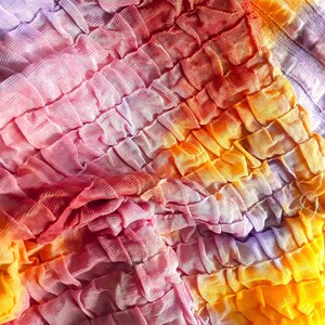 Może przedstawiać: Zbliżenie na tkaninę z wzorem tie-dye w odcieniach r&oacute;żowego, pomarańczowego i fioletowego. Tkanina jest zebrana i zmarszczona, tworząc fakturowaną powierzchnię.