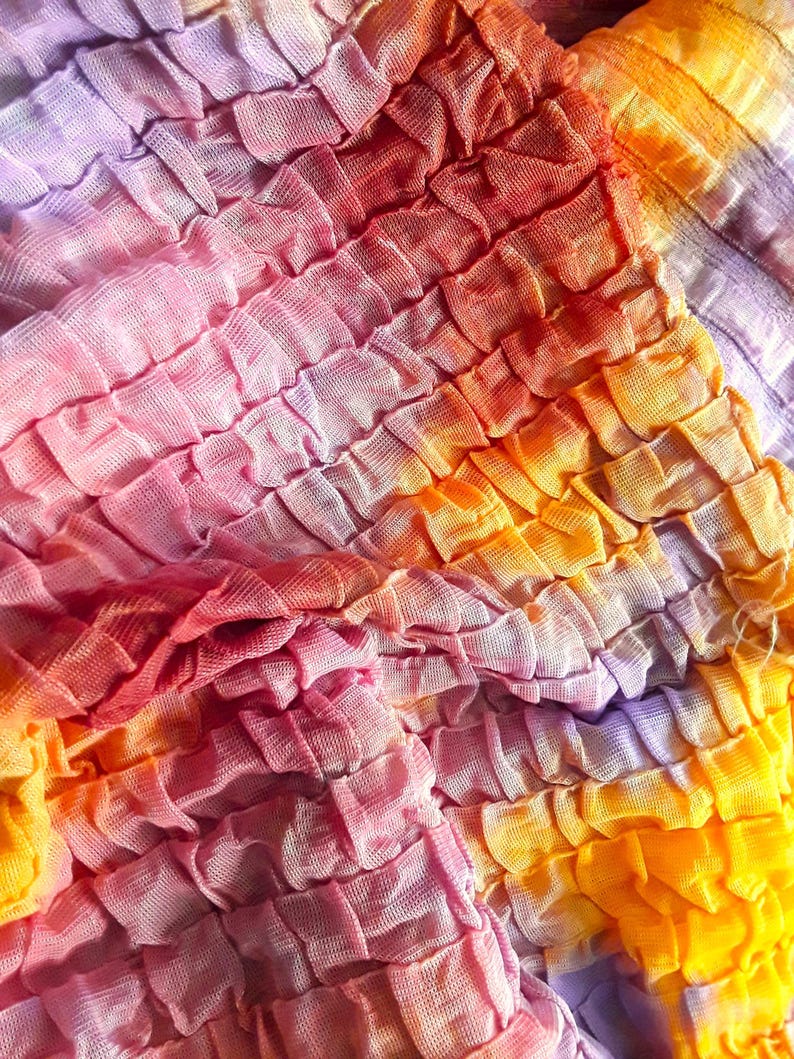 Może przedstawiać: Zbliżenie na tkaninę z wzorem tie-dye w odcieniach r&oacute;żowego, pomarańczowego i fioletowego. Tkanina jest zebrana i zmarszczona, tworząc fakturowaną powierzchnię.