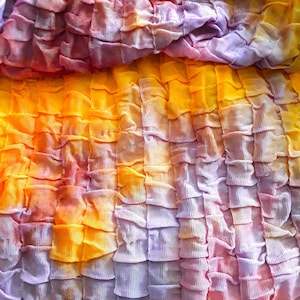 Może przedstawiać: Zbliżenie na tkaninę z wzorem tie-dye w odcieniach fioletu, pomarańczy i ż&oacute;łci. Tkanina jest zebrana i zmarszczona, tworząc fakturowany wygląd.