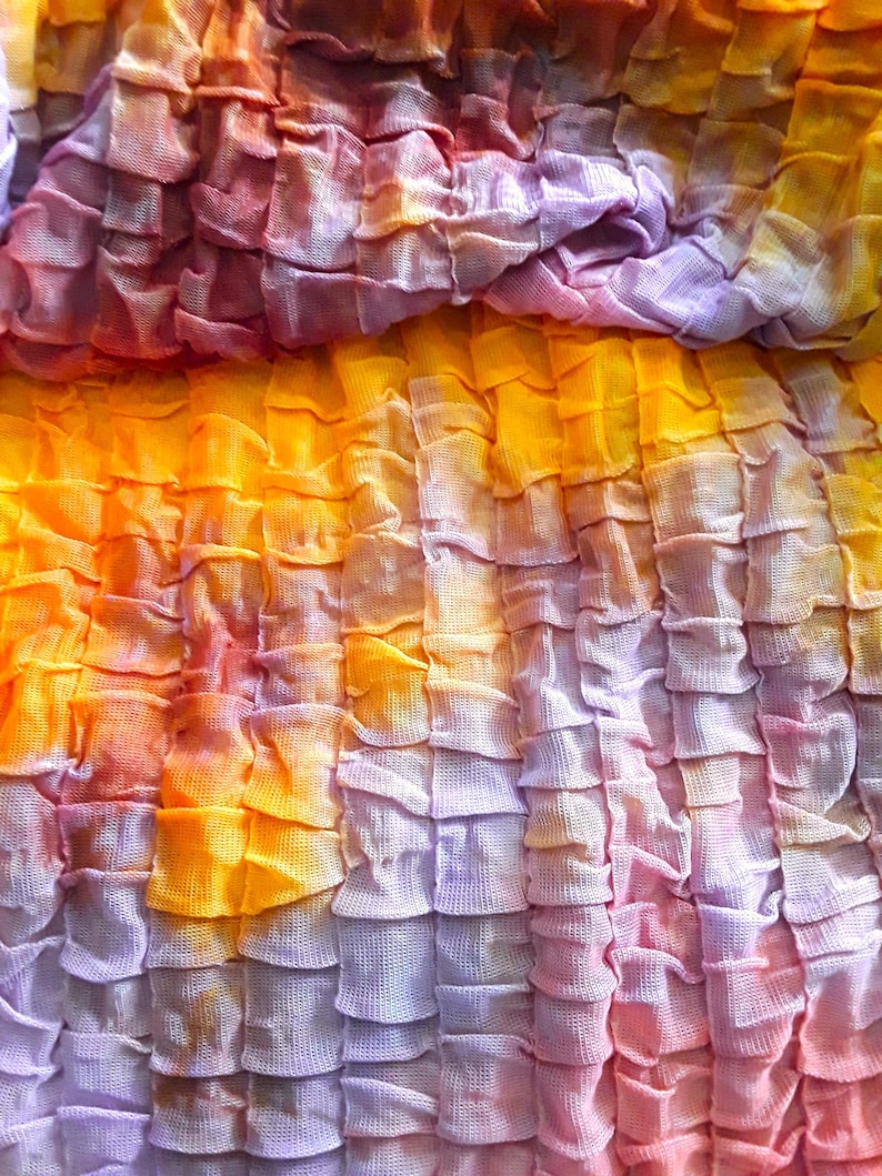 Może przedstawiać: Zbliżenie na tkaninę z wzorem tie-dye w odcieniach fioletu, pomarańczy i ż&oacute;łci. Tkanina jest zebrana i zmarszczona, tworząc fakturowany wygląd.