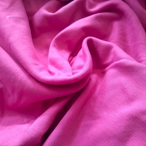 May include: Close-up of a vibrant pink fabric with soft folds and a textured surface. The material appears to be a knit or jersey, with a smooth, slightly shiny appearance. The color is a bright, saturated pink.