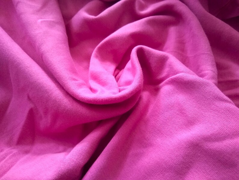 May include: Close-up of a vibrant pink fabric with soft folds and a textured surface. The material appears to be a knit or jersey, with a smooth, slightly shiny appearance. The color is a bright, saturated pink.