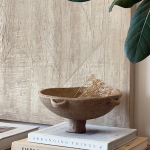 Könnte beinhalten: Eine beige, texturierte Keramikschale auf einem Sockel mit zwei kleinen seitlichen Griffen, die einen kleinen Zweig getrockneter Blumen enthält. Die Schale steht auf einem Stapel aus zwei Büchern mit den Titeln Arranging Things und Eight Homes Clements Design.