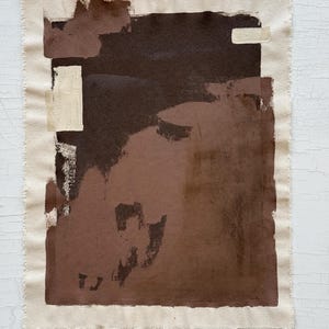 May include: Abstract artwork on a canvas with a raw edge. The painting features a dark brown central shape with lighter brown and beige accents. The canvas is rectangular and mounted on a white surface.