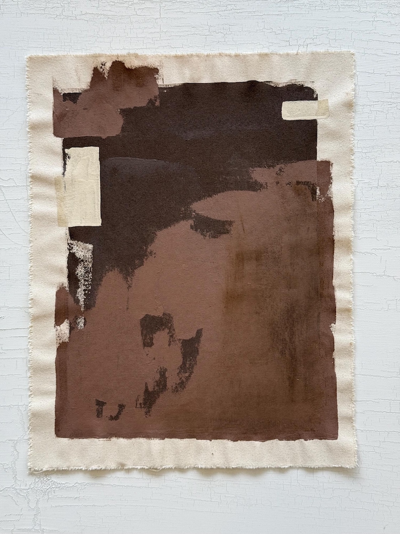May include: Abstract artwork on a canvas with a raw edge. The painting features a dark brown central shape with lighter brown and beige accents. The canvas is rectangular and mounted on a white surface.