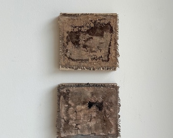 May include: Two square, abstract artworks. Each piece features a textured, brown surface with a darker central design and a frayed edge. The artworks are mounted on a white wall, showcasing a minimalist aesthetic.