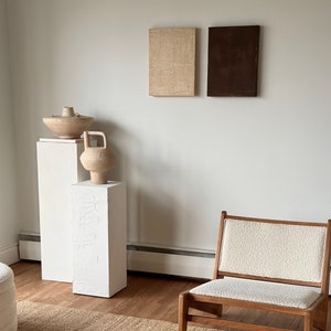 May include: Two abstract paintings, one beige and one brown, hang on a white wall above two white pedestals. A ceramic vase and bowl sit on the pedestals. A wooden chair with a white cushion sits in front of a woven rug.