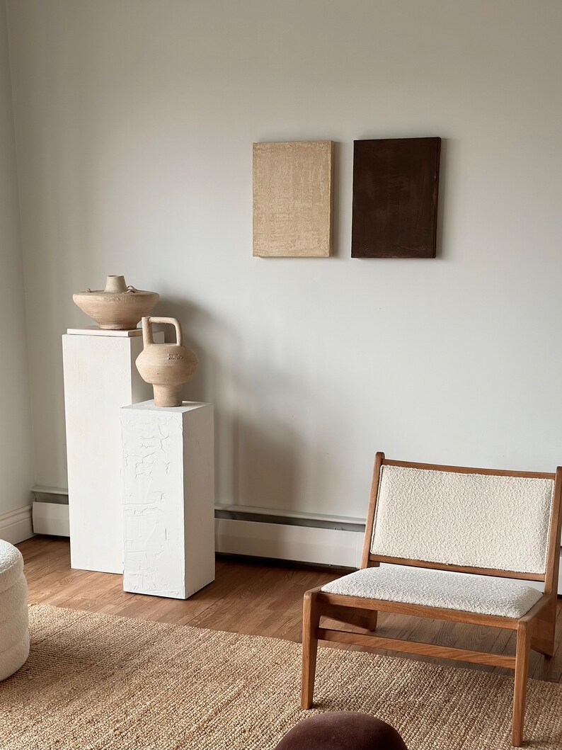 May include: Two abstract paintings, one beige and one brown, hang on a white wall above two white pedestals. A ceramic vase and bowl sit on the pedestals. A wooden chair with a white cushion sits in front of a woven rug.