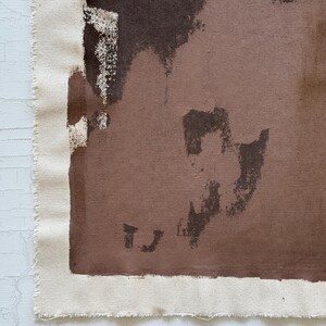 May include: Abstract art piece featuring a canvas with a raw edge. The artwork uses a palette of brown and beige tones, with brushstrokes and shapes creating a textured effect. The canvas is mounted on a white surface.