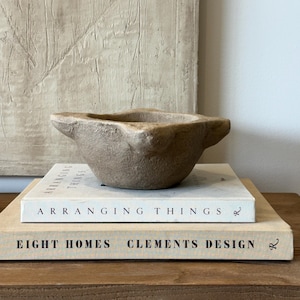 以下が含まれることがあります： Arranging ThingsとEight Homes Clements Designというタイトルの2冊の本の上に置かれた、4つの突起のあるハンドルが付いた素朴なベージュ色の石製乳鉢。乳鉢は質感のある風化した表面をしており、開いたボウル状の形をしています。