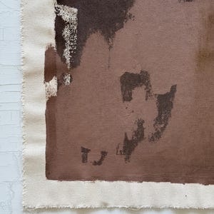 May include: Abstract art piece featuring a canvas with a raw edge. The artwork uses a palette of brown and beige tones, with brushstrokes and shapes creating a textured effect. The canvas is mounted on a white surface.