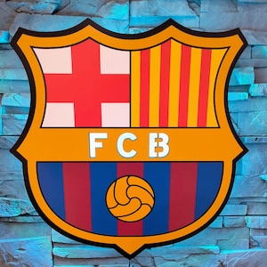 Può includere: Uno scudo giallo e blu con il logo del FC Barcelona. Il logo presenta una croce rossa e bianca, strisce verticali gialle e rosse, un pallone da calcio e le lettere FCB.