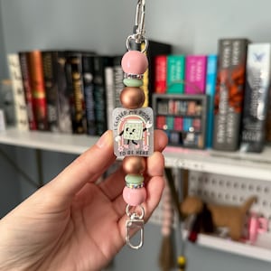 May include: A beaded keychain with a silver clasp. The keychain features pink, green, and copper-colored beads. A rectangular charm displays a book with a face, a rainbow, and the text "I CLOSED MY BOOK TO BE HERE."