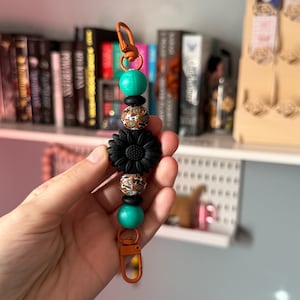 May include: A beaded keychain with a black sunflower, teal beads, and patterned beads. The keychain has a bronze-colored clasp and connector rings. The beads are strung together with a bronze-colored wire.
