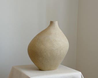 Puede incluir: Un jarrón de cerámica beige asimétrico con un acabado mate y texturizado. El recipiente presenta un cuerpo redondeado y bulboso que se estrecha hacia arriba en un cuello cilíndrico estrecho con una pequeña abertura circular en la parte superior.