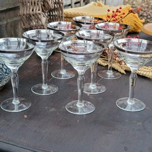 May include: Set of eight clear glass champagne coupes with silver rims. The glasses are arranged on a dark wooden surface.