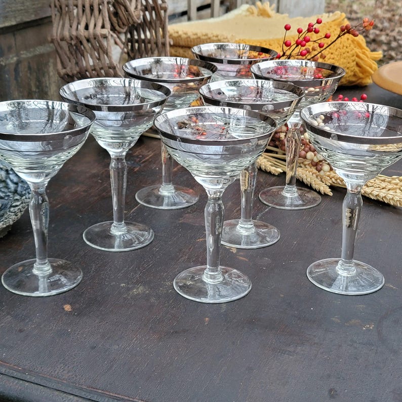 May include: Set of eight clear glass champagne coupes with silver rims. The glasses are arranged on a dark wooden surface.