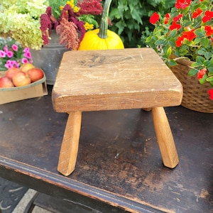 May include: A small, square wooden stool with three angled legs. The stool is made of light brown wood with visible grain and wear marks. The legs are tapered and attached to the underside of the seat. The stool is likely vintage or antique.