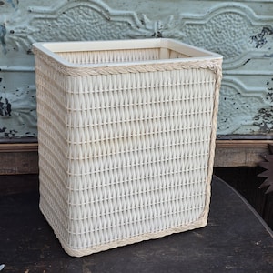 May include: A rectangular, off-white wicker basket with a braided trim around the top and base. The basket has a woven pattern and is designed for storage. The basket is made of a light-colored material.