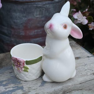 May include: A white ceramic rabbit figurine standing next to a small, round basket-weave planter. The rabbit has pink-tinted ears and nose with brown eyes. The planter features a green rim and a painted floral design with pink flowers and green leaves.