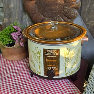 May include: Vintage Crock Watcher slow cooker with a glass lid and brown accents. The off-white body features a wheat-like design. The control panel reads "Crock Watcher by Hamilton Beach". The slow cooker sits on a red and white checkered surface.