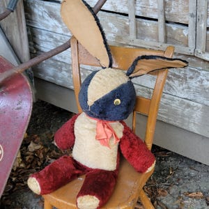 May include: Vintage plush bunny rabbit toy with a cream-colored body, red arms and legs, and a navy blue and cream-colored head. The bunny has long ears, a red ribbon, and is seated on a wooden chair. The toy has a classic, antique appearance.