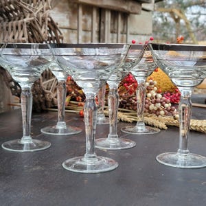 May include: Set of six clear glass champagne coupes with silver rims. The glasses are arranged on a dark wooden surface.