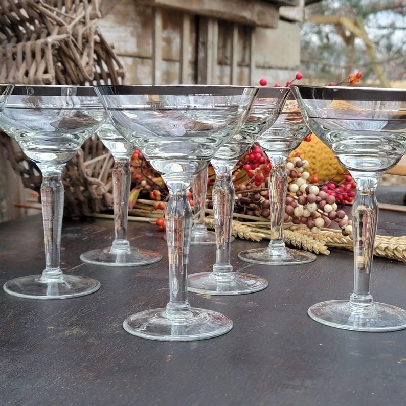 May include: Set of six clear glass champagne coupes with silver rims. The glasses are arranged on a dark wooden surface.