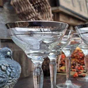 May include: Set of four clear glass cocktail glasses with a silver rim. The glasses are arranged in a row, with the front glass in focus. The glasses are on a table with a blurry background.
