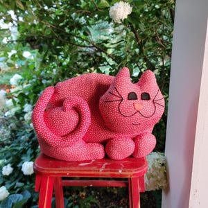 May include: A plush, red cat with a white floral pattern sits on a small red stool. The cat has black eyes, whiskers, and a pink nose. Its tail curls into a heart shape. The background features greenery and white flowers.