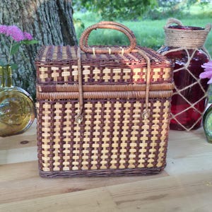 May include: A rectangular woven basket featuring a two-tone pattern of light tan and dark brown reeds. The basket includes a hinged lid with a central arched handle and two vertical straps that secure the lid to the main body of the container.