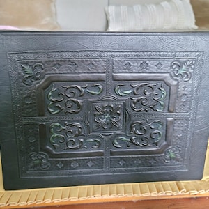 May include: A rectangular dark brown box with a textured faux leather finish. The lid features an embossed design with intricate scrollwork, floral motifs, and geometric borders, accented with subtle hints of green within the recessed areas.