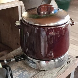 May include: A dark maroon ceramic electric pot with a shiny chrome base and a matching maroon lid featuring a brown knob. The appliance includes a black power cord with a metal spring strain relief attached to the side of the chrome base.