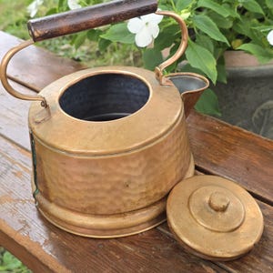 May include: A hammered copper kettle with a dark brown wooden handle and a matching circular lid. The kettle features a wide opening at the top, a curved spout, and a metallic finish with visible texture across the exterior surface.