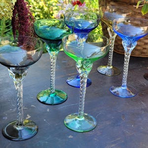 May include: A collection of colorful, stemmed champagne glasses. Each glass features a twisted stem and a bowl in shades of blue, green, and yellow. The glasses are arranged on a dark surface, with a blurred background of greenery.