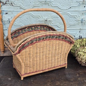 May include: A woven wicker basket with a curved handle. The basket features a rectangular base and is adorned with woven details in shades of tan, green, and burgundy. The handle is arched and made of the same woven material.
