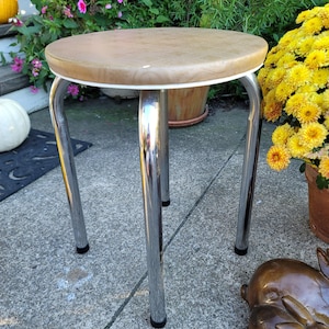 May include: A vintage stool with a round, light brown seat and chrome legs. The seat has a white trim. The legs are curved and end with black rubber feet. The stool is outdoors, near potted plants and a decorative pumpkin.