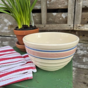 May include: A cream-colored ceramic mixing bowl with a ribbed exterior featuring a wide horizontal band in dusty pink bordered by two thin blue stripes. The bowl has a smooth interior finish and a rounded rim, showcasing a classic kitchen design.